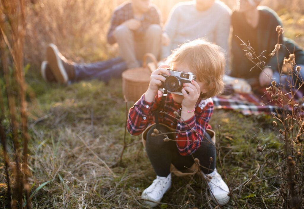 Small girl with family on picnic in autumn nature, taking photographs with camera